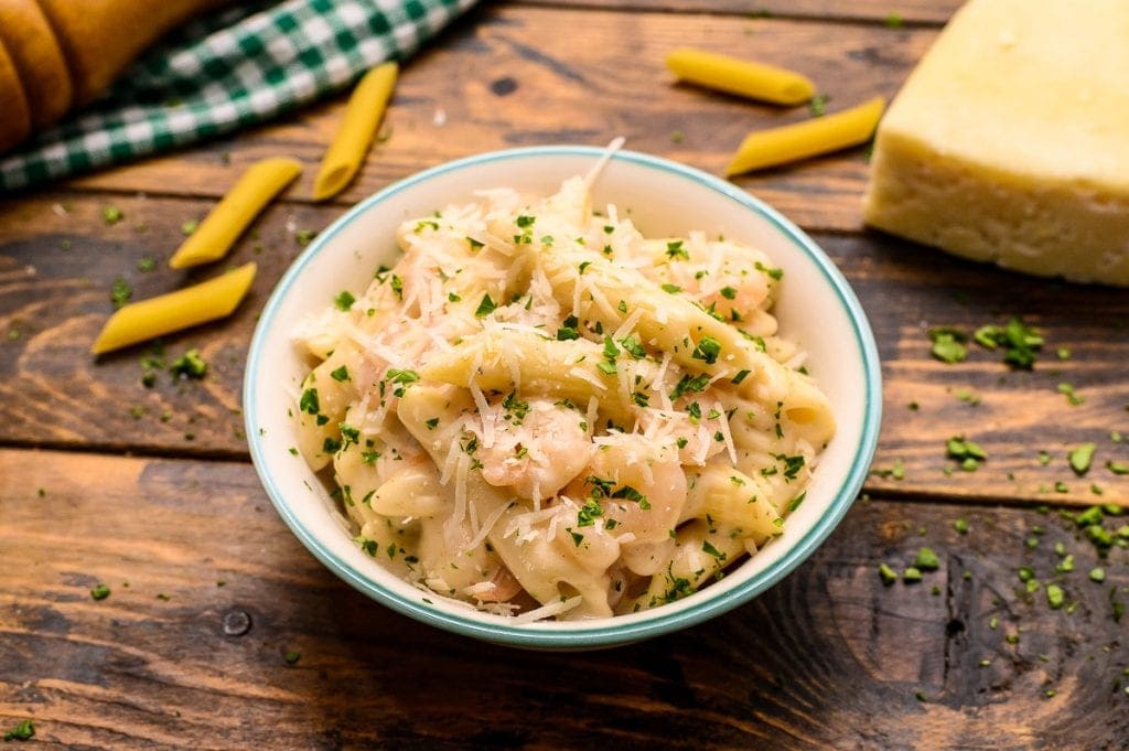 Bowl of shrimp penne pasta on a brown background with dry noodles and cheese in background