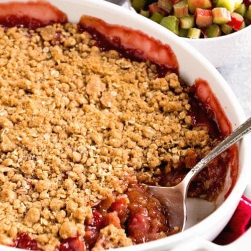 White casserole pan of strawberry rhubarb crisp next to two small bowls of strawberries and rhubarb