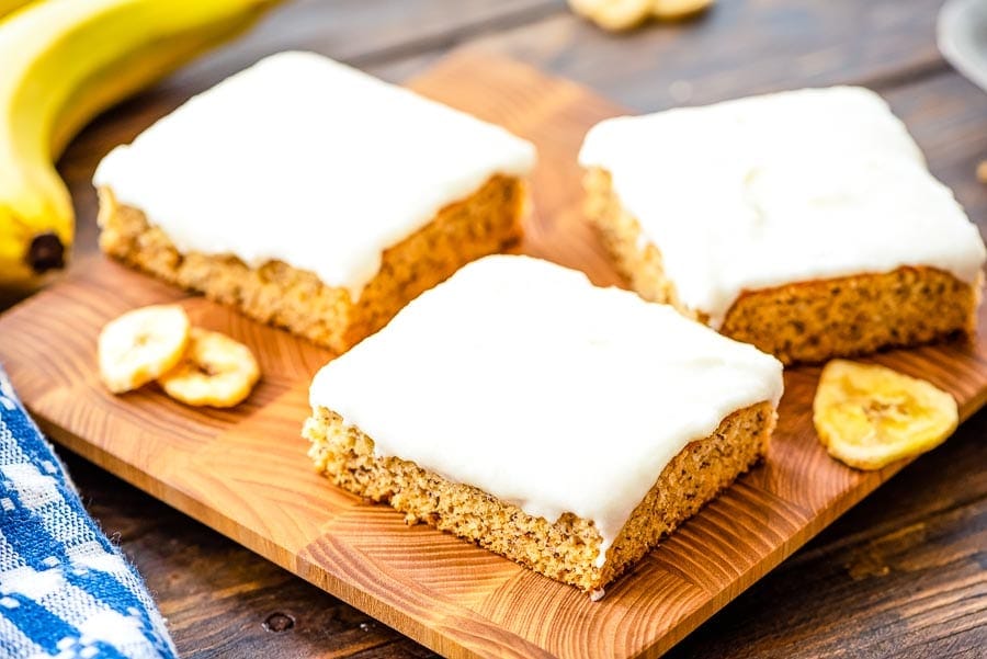 A wooden cutting board with three slices of cream cheese frosted banana bars on it