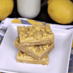 Three lemon crumb bar stacked on a white plate in front of two lemons and a glass of milk