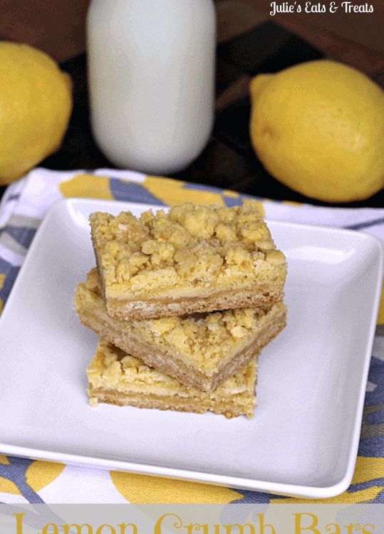 Three lemon crumb bar stacked on a white plate in front of two lemons and a glass of milk