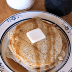 A short stack of zucchini bread pancakes topped with a pad of butter and syrup on a white plate in front of a glass of milk and a mug of coffee