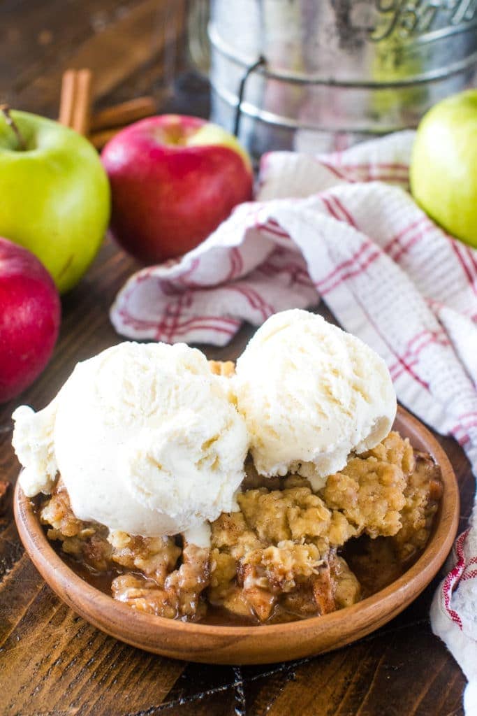 Easy Apple Crisp Recipe on brown plate topped with two scoops of vanilla ice cream. In the background a white and red towel, green and red apples and an old metal sifter.