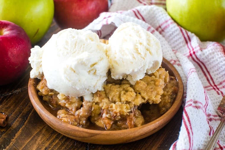 Apple Crisp on brown plate topped with two scoops of vanilla ice cream with apples and white and red striped towel in background