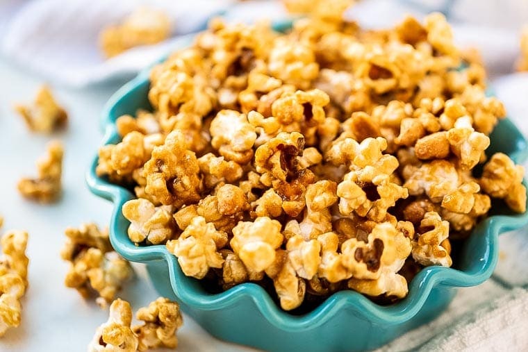 Teal bowl with scalloped edges filled wit homemade caramel corn with it scattered next to it.