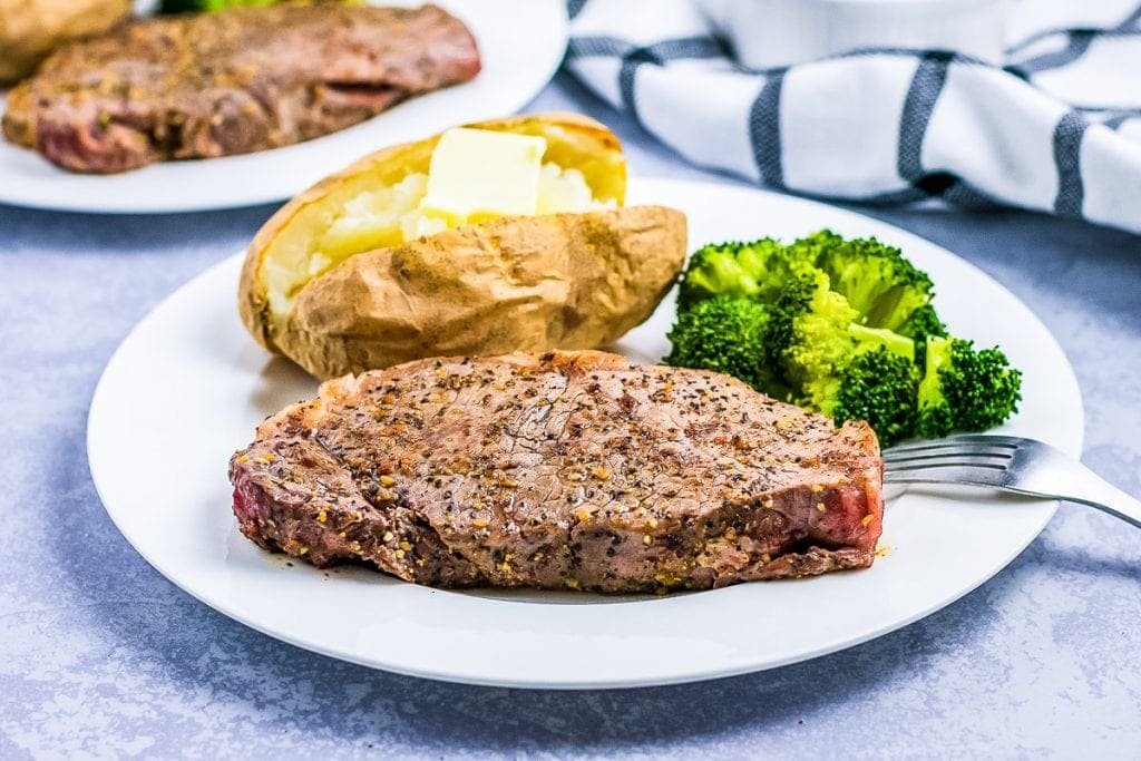 Seasoned steak on a white plate with baked potato and broccoli