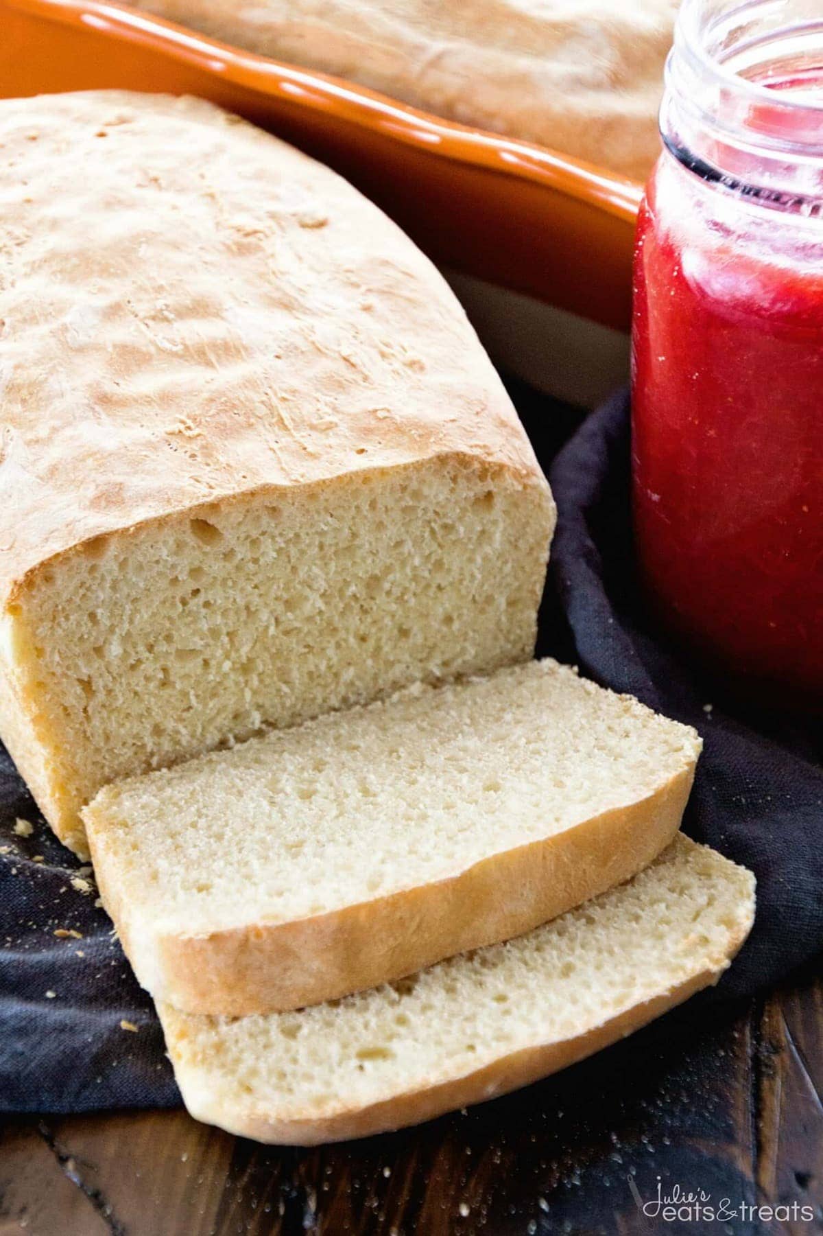 Loaf of sliced english muffin bread with jam behind it in glass jar and another loaf of bread in pan