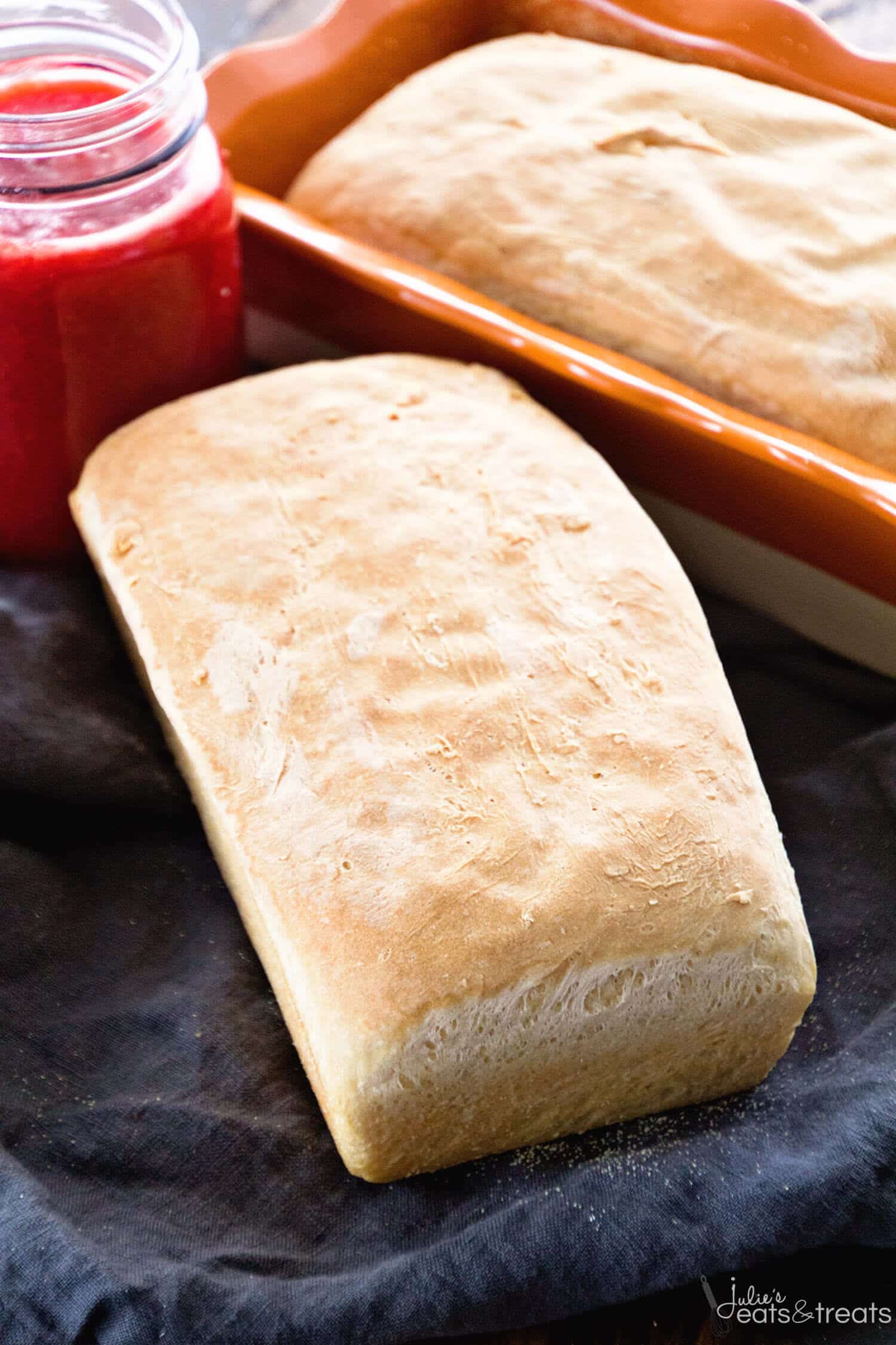 Whole loaf of bread on dark blue napkin with another loaf in pan behind it and a jar of jam in background