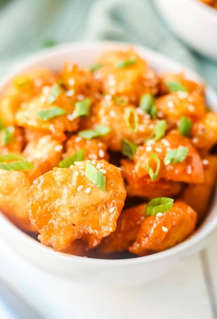 White bowl with sweet and sour chicken pieces topped with sesame seeds and slice green onions