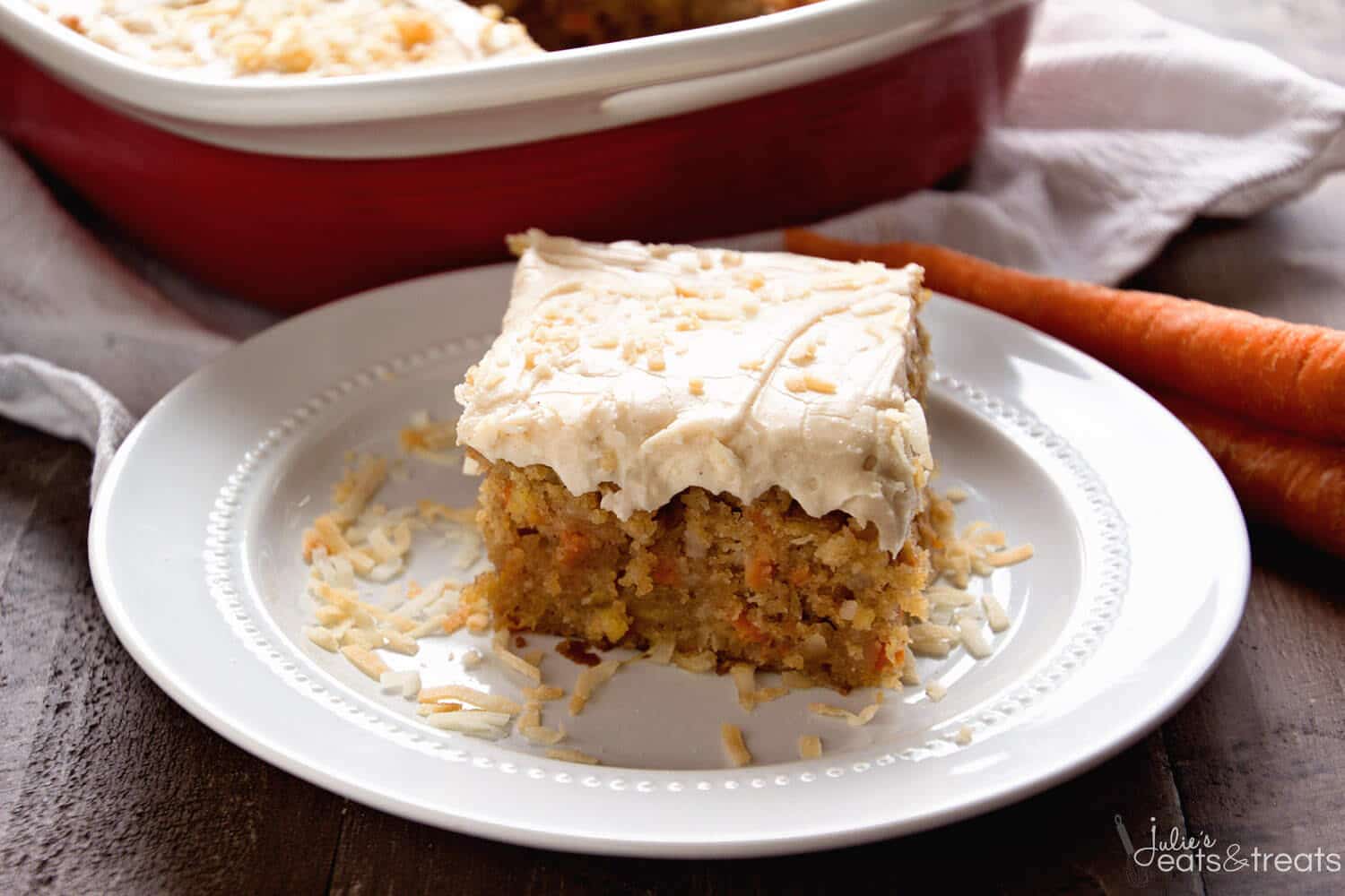 White plate with carrot poke cake piece on it with pan of cake behind it and carrots next to it