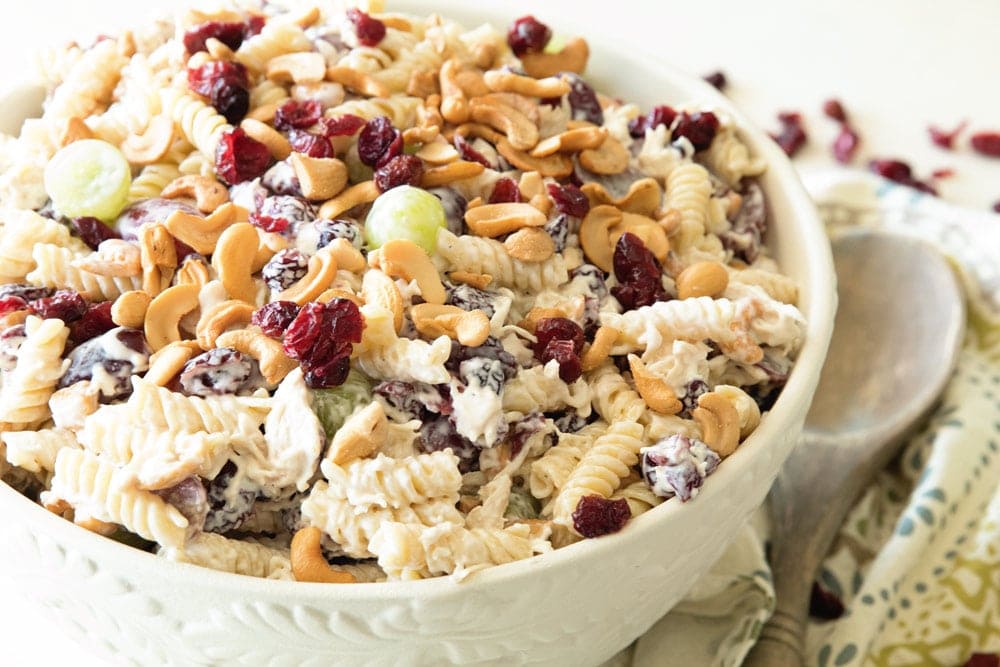 Closeup image of prepared cashew chicken rotini salad and cashews on top of it and a wooden spoon and napkin beside it