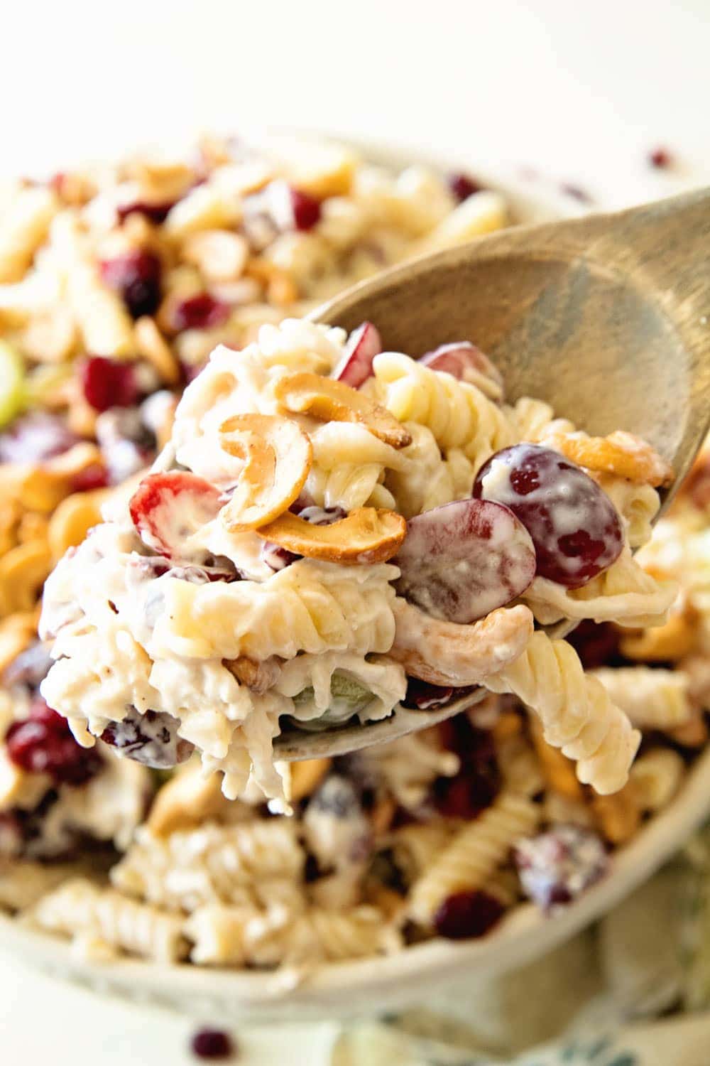 Wooden spoon with cashew chicken rotini salad with grapes in it and a bowl of salad in background