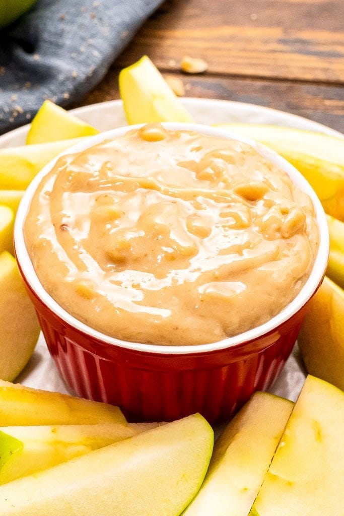 A red bowl on a white plate full of apple brickle dip. Sliced apples arranged on plate around the bowl.