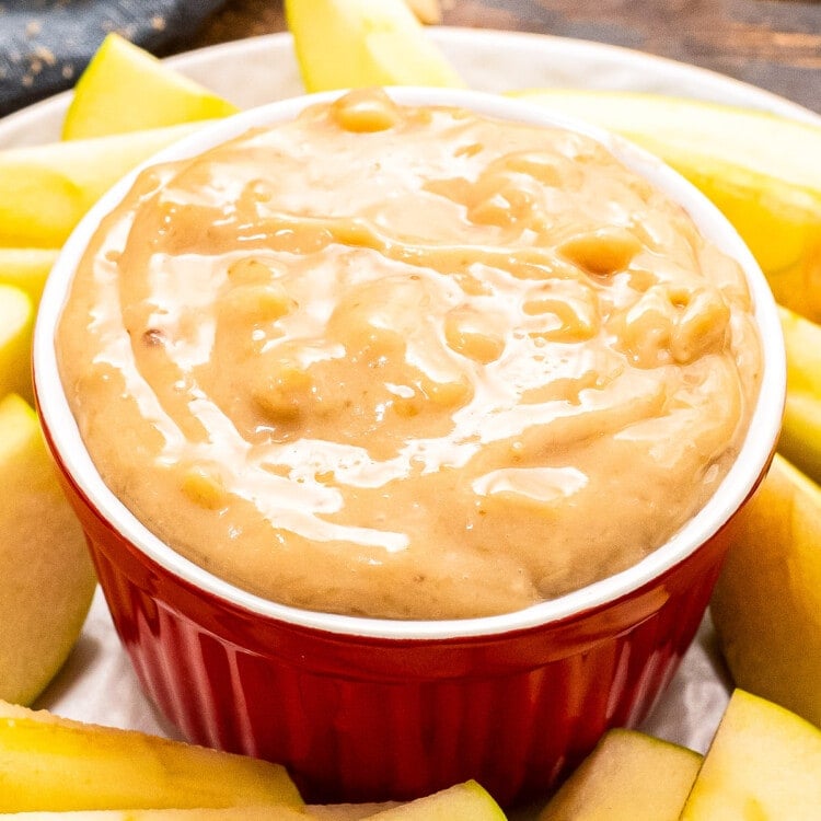 A red bowl on a white plate full of apple brickle dip. Sliced apples arranged on plate around the bowl.