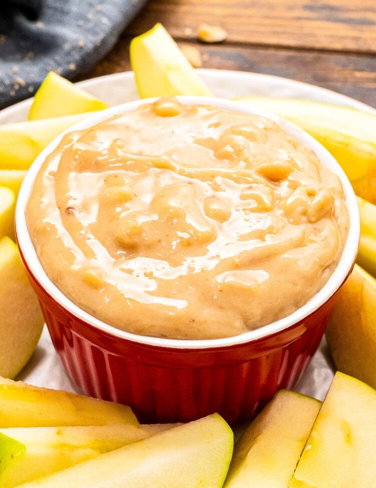 A red bowl on a white plate full of apple brickle dip. Sliced apples arranged on plate around the bowl.