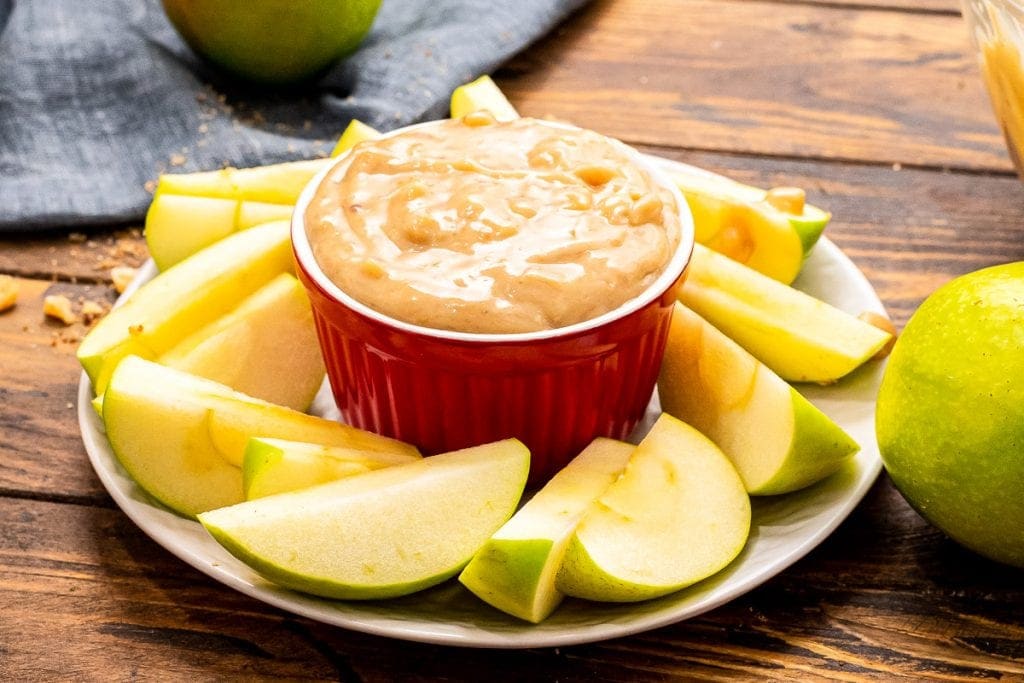 A round plate with slices of apples around a red bowl of apple brickle dip.