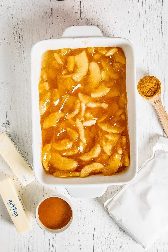 Apple Pie filling in white baking dish with butter sticks, cinnamon and caramel beside it.