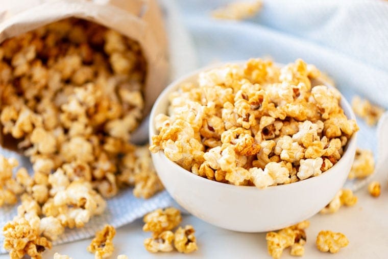 Caramel corn in white bowl with a bag in the background full of more and it spilled on the surface.