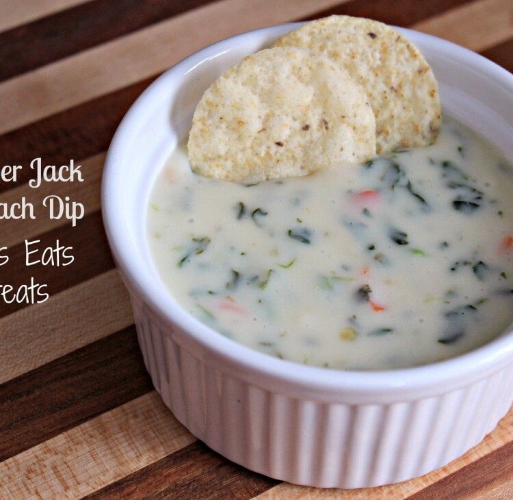 A white bowl of pepper jack spinach dip with two chips in it sitting on a wood board