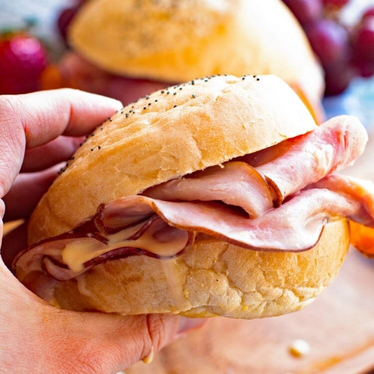 Hot Ham Cheese Sandwich in Hand over a wood plate with another sandwich and grapes on it