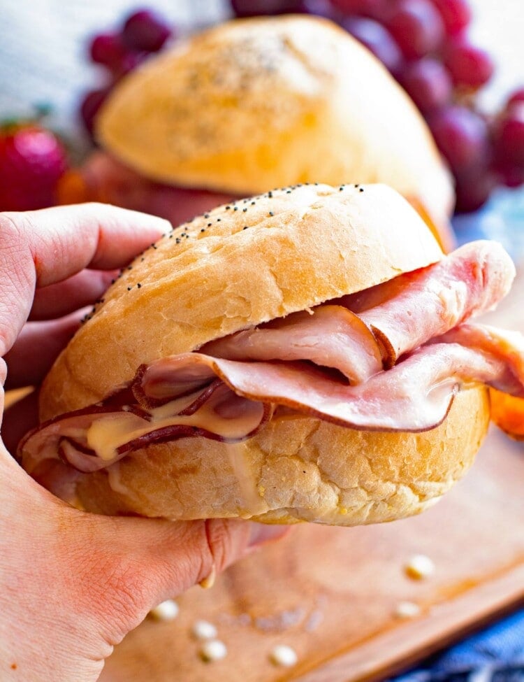 Hot Ham Cheese Sandwich in Hand over a wood plate with another sandwich and grapes on it