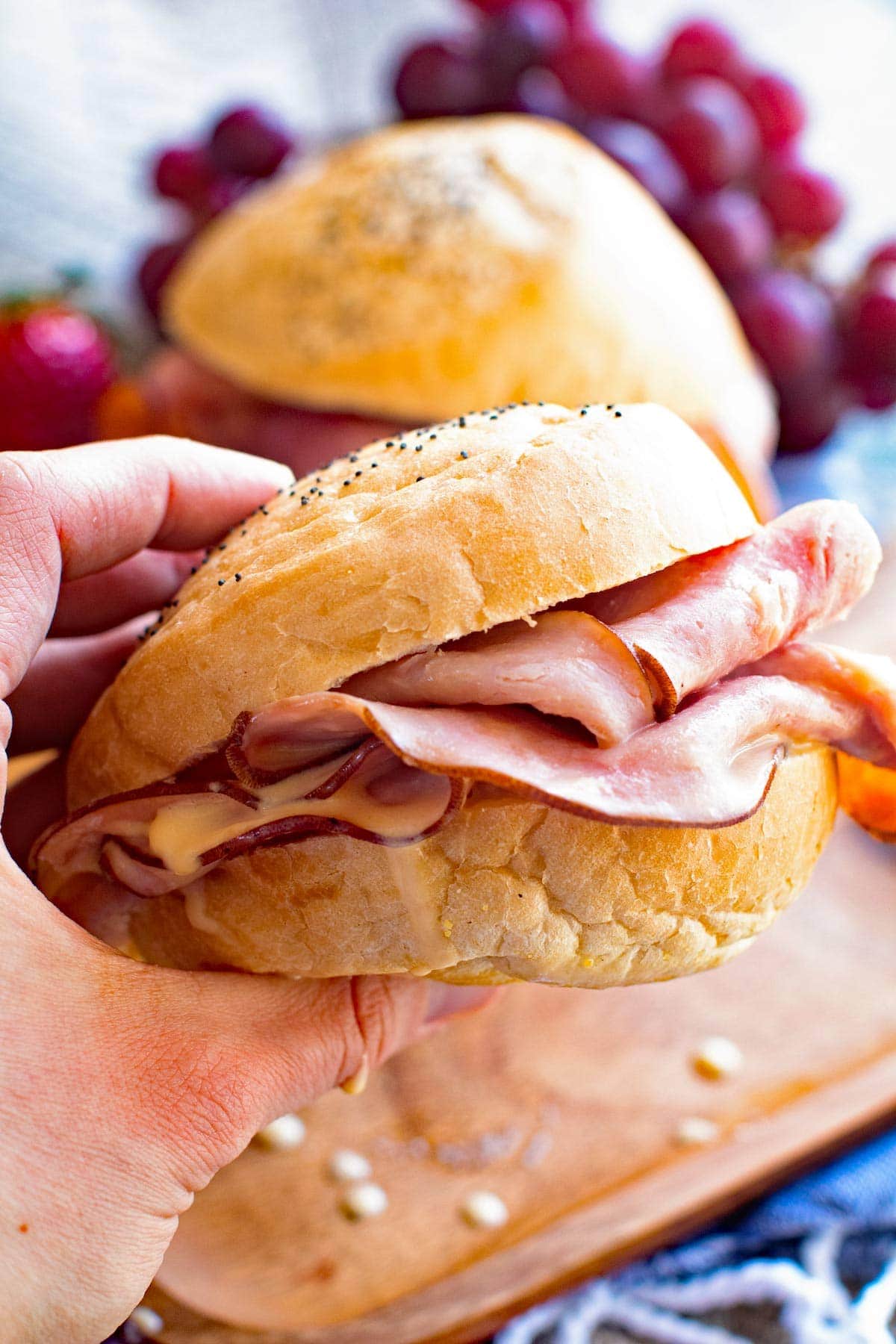 Hot Ham Cheese Sandwich in Hand over a wood plate with another sandwich and grapes on it