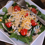 A square white bowl with spinach, tomatoes, dressing, shredded cheese, and a fork sitting on a white square plate