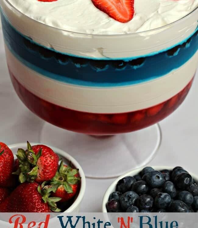A tall clear glass bowl of red, white and blue salad with two bowls of strawberries and blueberries in front of it