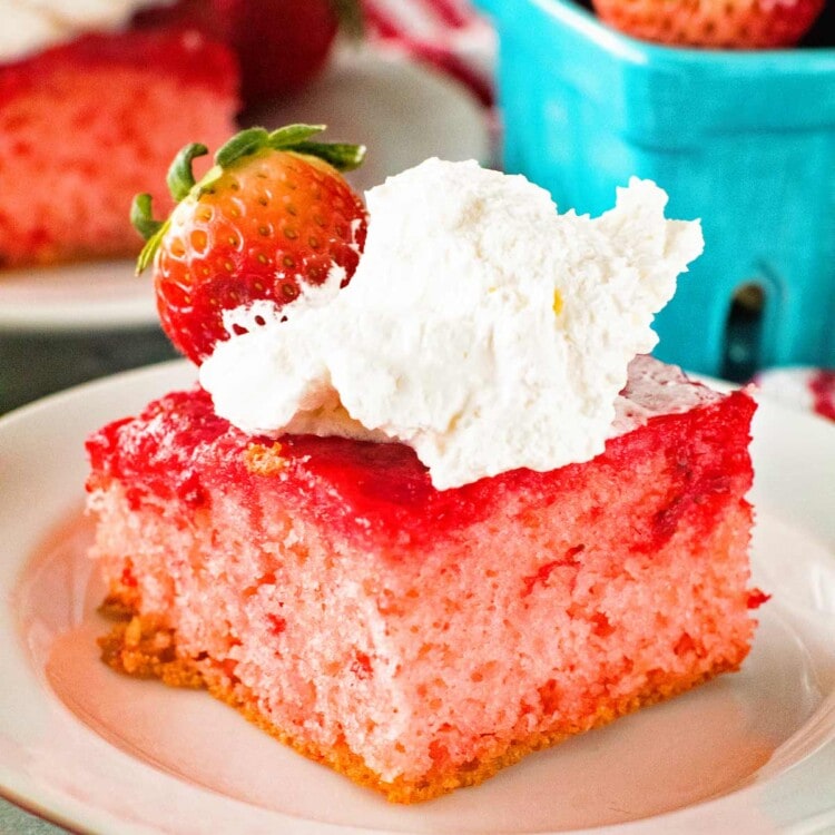 A piece of Upside Down Strawberry Cake topped with whipped cream and a strawberry sitting on a white plate in front of a blue carton of strawberries
