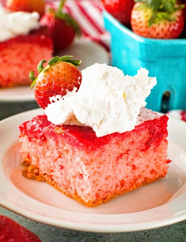 A piece of Upside Down Strawberry Cake topped with whipped cream and a strawberry sitting on a white plate in front of a blue carton of strawberries