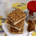 Three biscoff krispie mallow bars stacked on a white cloth napkin next to a spoon full of biscoff spread, a jar of biscoff spread, and a glass of milk