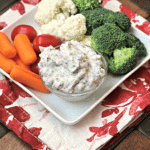 A square white plate with carrots, cherry tomatoes, cauliflower, broccoli, and a bowl of dill veggie dip on it