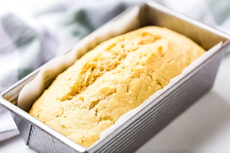 Loaf of beer bread in a loaf pan that is lined with parchment paper. A blue and white checkered cloth napkin in background.