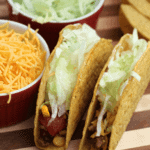 Two Texas tacos on a wood board next to two bowls containing shredded cheese and shredded lettuce