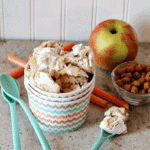 A bowl of apple cinnamon ice cream next to blue spoons, an apple, and a bowl of cinnamon chips