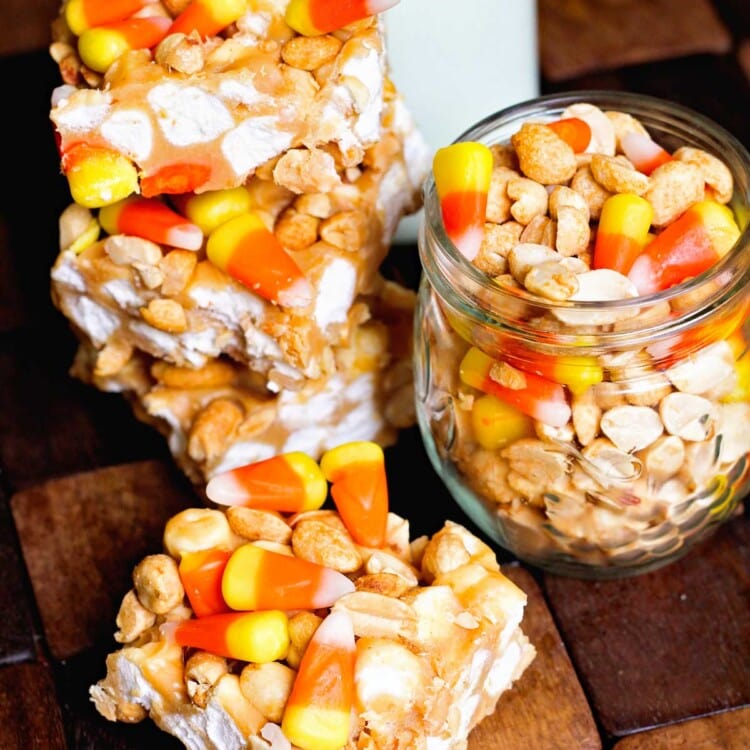 Four candy corn peanut bars on a wood board with a jar of candy corn and peanuts and a jar of milk