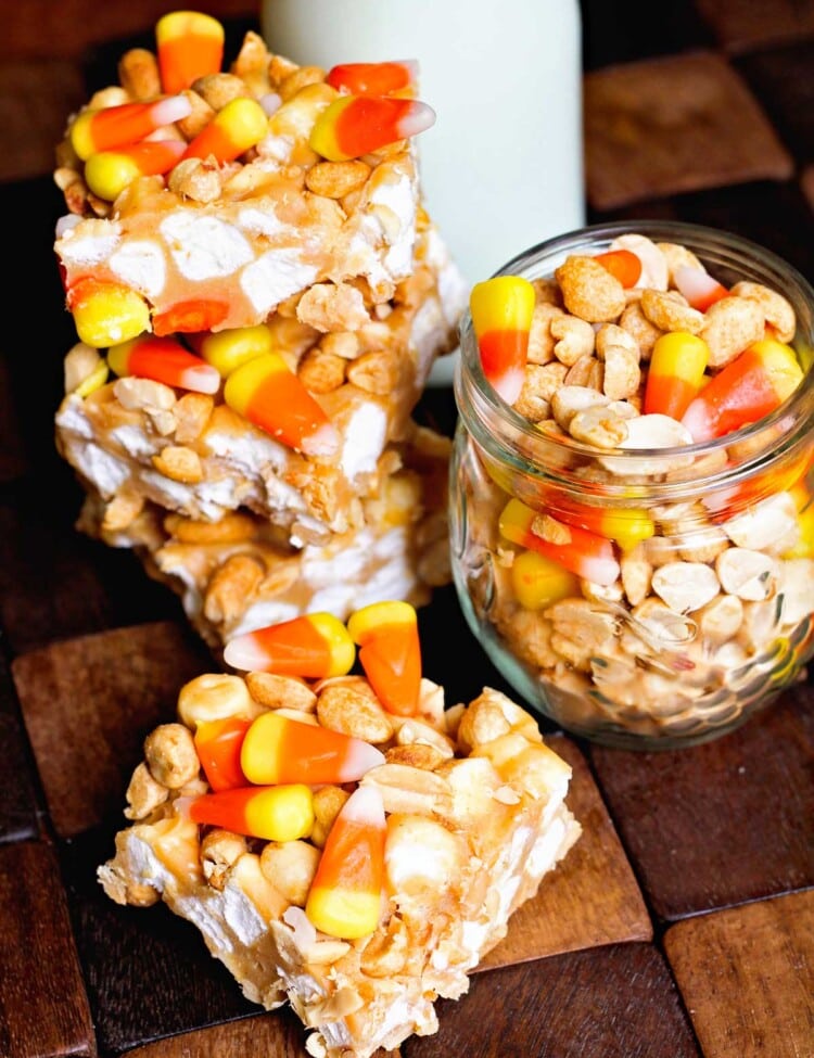 Four candy corn peanut bars on a wood board with a jar of candy corn and peanuts and a jar of milk