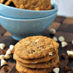 Stack of four cinnamon chip pumpkin cookies on a wood board scattered with cinnamon chips and white chocolate chips and a blue bowl of cookies in the background