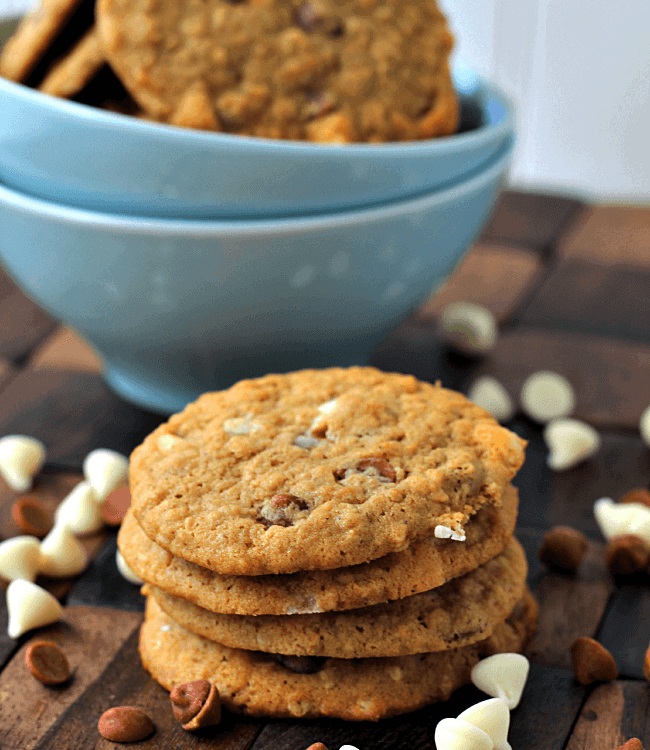 Stack of four cinnamon chip pumpkin cookies on a wood board scattered with cinnamon chips and white chocolate chips and a blue bowl of cookies in the background