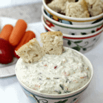 A bowl of garden vegetable spread with two pieces of bread in it and a bowl of bread behind it