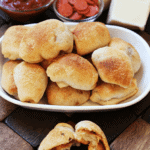 Homemade pepperoni pizza rolls on a white plate in front of a bowl of marinara sauce, a bowl of pepperoni, and some cheese