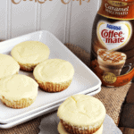 Two caramel macchiato sugar cookie cups stacked on wax paper next to a square white plate with four cups on it and a bottle of caramel macchiato coffee creamer