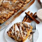 Piece of overnight pumpkin cheesecake and a fork on a white plate in front of a glass dish of the rest of the casserole