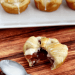 Apple cranberry cheesecake pie bite on a table with a bite out of it next to a spoon with icing on it and in front of a white plate with three more pie bites on it