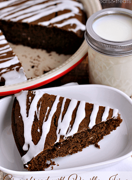 Slice of gingerbread with vanilla icing in a white bowl in front of a glass of milk and a pie plate containing the rest of the gingerbread