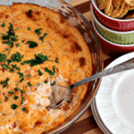 Glass pie plate of cheesy bruschetta dip with a spoon sitting on a wood cutting board with a stack of white plates and a bowl of corn chips