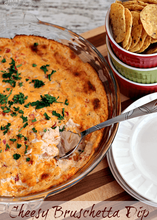 Glass pie plate of cheesy bruschetta dip with a spoon sitting on a wood cutting board with a stack of white plates and a bowl of corn chips