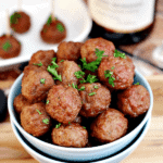 Two bowls stacked with the top full of cranberry pinot noir meatballs on a table in front of a white plate of meatballs with toothpicks in them and a bottle of wine