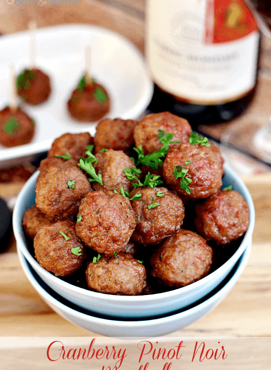 Two bowls stacked with the top full of cranberry pinot noir meatballs on a table in front of a white plate of meatballs with toothpicks in them and a bottle of wine