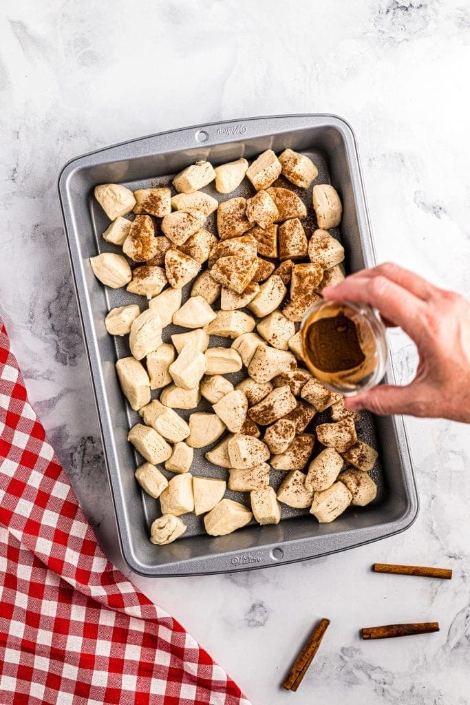 Biscuit pieces in cake pan being sprinkled with cinnamon